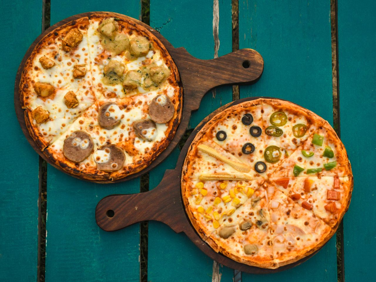 services-03 Two pizzas with various toppings on wooden boards over a rustic green table, captured in a top-down view.