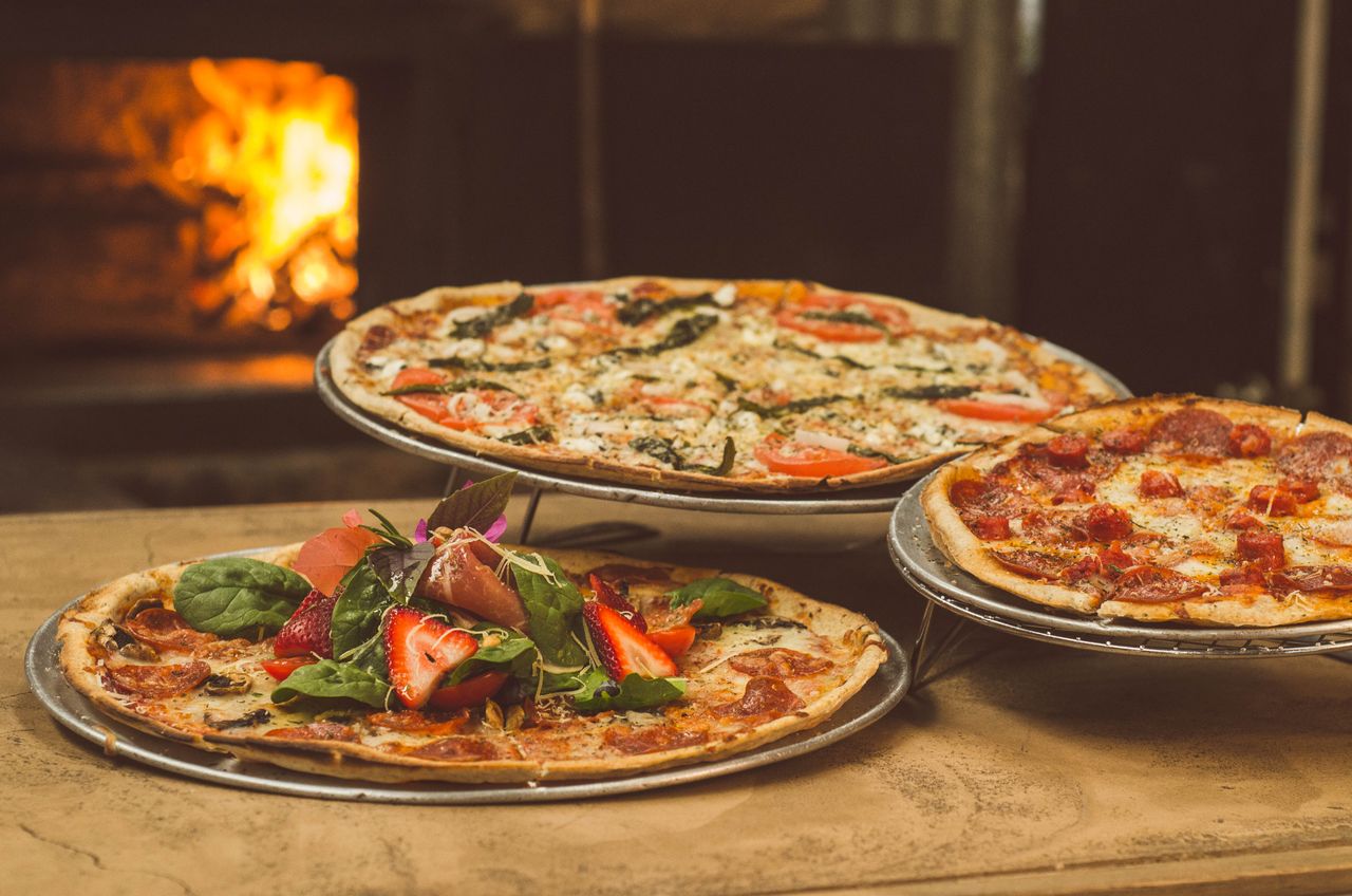 about-us Trio of gourmet pizzas with unique toppings near a wood-fired oven, ideal for food enthusiasts.